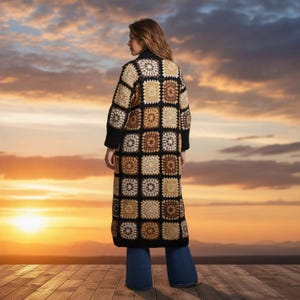 May include: A long, open-front cardigan featuring a patchwork of crocheted granny squares in shades of cream, beige, and brown, bordered by black yarn. The garment has long sleeves and a straight hem, worn over blue flared denim pants.