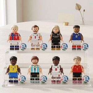 May include: Eight miniature plastic soccer player figures arranged on a two-tiered clear acrylic display stand. Each figure wears a different national or club team jersey with a corresponding number and is accompanied by a small soccer ball.
