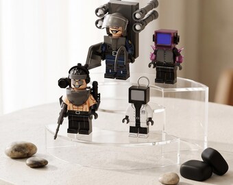 May include: Four plastic minifigures displayed on a tiered clear acrylic stand. One figure wears a tactical helmet and backpack, another has a purple television head, one is shirtless with a headset, and the last wears a white lab coat with a gray screen head.