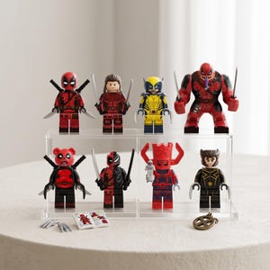 May include: A collection of eight superhero-themed plastic minifigures displayed on a two-tiered clear acrylic stand. The figures feature various character designs including red and black suits, yellow and blue armor, and a large red monster figure.