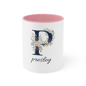 Puede incluir: Una taza de cerámica blanca con interior y borde rosa. El frente presenta una letra P grande de color azul marino oscuro decorada con flores y hojas verdes estilo acuarela. Debajo de la letra, el nombre presley está impreso en fuente cursiva negra.
