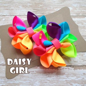 May include: Two rainbow-colored hair bows with a daisy-shaped design. The bows are made of multiple layers of ribbon in shades of pink, purple, orange, yellow, and green. The bows are arranged on a brown background with the text "DAISY GIRL" printed below.