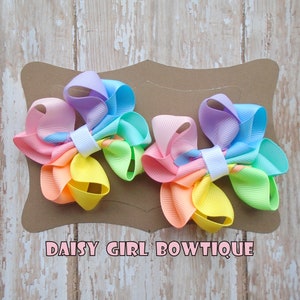 May include: Two pastel rainbow colored hair bows with white centers. The bows are on a brown card with the text "DAISY GIRL BOWTIQUE".