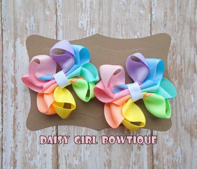 May include: Two pastel rainbow colored hair bows with white centers. The bows are on a brown card with the text "DAISY GIRL BOWTIQUE".
