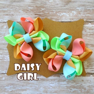 May include: Two colorful hair bows with a layered design in shades of green, orange, and pink. The bows are displayed on a brown card with the text "DAISY GIRL".