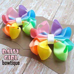 May include: Two rainbow-colored hair bows with a white center. The bows are made of a soft, textured fabric and are arranged on a brown background. The text "DAISY GIRL bowtique" is printed on the background.