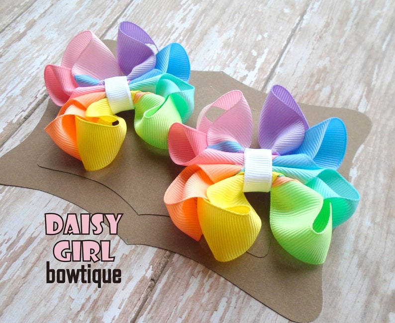 May include: Two rainbow-colored hair bows with a white center. The bows are made of a soft, textured fabric and are arranged on a brown background. The text "DAISY GIRL bowtique" is printed on the background.