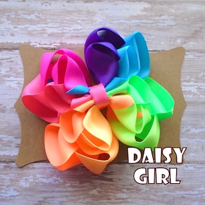 May include: A rainbow-colored hair bow with a layered, ruffled design. The bow is on a brown card with the text "DAISY GIRL" in white.