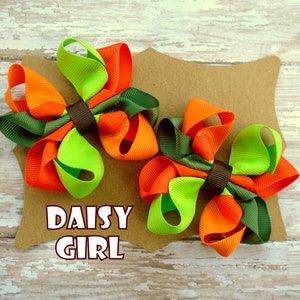 May include: Two green, orange, and brown hair bows with a brown center. The bows are on a brown card with the text "DAISY GIRL" printed on it.