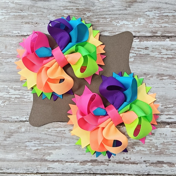 May include: Three vibrant hair bows in a rainbow of colours, featuring pink, orange, yellow, green, and purple. Each bow is layered and has a spiky, starburst-like base. The bows are set against a neutral brown background.