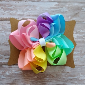 May include: A multi-colored bow made of ribbon in shades of pink, yellow, green, blue, and purple. The bow is tied with a white ribbon in the center.