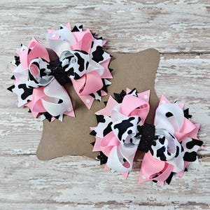 May include: Two handmade hair bows with a cow print design. The bows feature layers of pink, white, and black and white patterned ribbons, with a black glitter center. The bows are attached to a brown card.