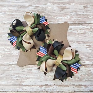 May include: Two handmade hair bows with a patriotic theme. Each bow features layers of tan, olive green, brown, and black ribbons, accented with small American flag details and a gold glitter center. The bows are displayed on a brown cardboard backing.