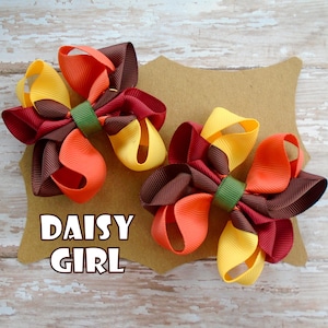 May include: Two brown, orange, and yellow hair bows with green centers. The bows are on a brown card with the text "DAISY GIRL" in white.