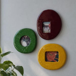 Pode incluir: Três molduras circulares e brilhantes para parede nas cores verde, bordô escuro e amarelo vivo. Cada moldura apresenta uma forma orgânica, lisa e arredondada, com uma abertura retangular central que exibe uma pequena fotografia.