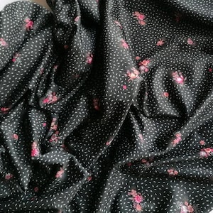 Op de afbeelding: Een zwarte stof met een herhalend patroon van kleine witte streepjes en trossen roze bloemen. Het materiaal is in plooien gedrapeerd, wat de gelijkmatige verdeling van de bloemen- en streepjesprint over het donkere oppervlak laat zien.