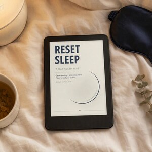 Könnte beinhalten: Ein schwarzer E-Reader mit dem Text RESET SLEEP 7-DAY SLEEP RESET, Calmer evenings + better sleep habits, 7 days to reset your routine, A simple wellness guide und einer Mondsichel-Grafik, platziert neben einer Tasse und einer Schlafmaske.