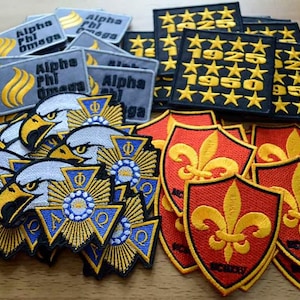 Pode incluir: Uma coleção de emblemas bordados incluindo emblemas retangulares cinzentos Alpha Phi Omega, emblemas quadrados pretos com estrelas douradas e o texto 1925 1950, emblemas de cabeças de águia com emblemas azuis e dourados e emblemas vermelhos em forma de escudo.