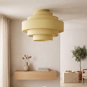 Peut inclure: Un plafonnier à plusieurs niveaux composé de cinq abat-jours circulaires concentriques en tissu de couleur jaune pâle. La structure s'affine du centre vers le haut et le bas, créant une silhouette géométrique superposée pour l'éclairage intérieur.