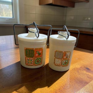 May include: Two white cylindrical plastic canisters with wire bail handles and lids. Each canister features a square graphic print on the front, displaying a grid of orange and green geometric and floral patterns on a white background.