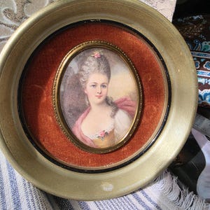 May include: An oval portrait miniature of a person with an elaborate hairstyle, wearing a pink shawl and a light-colored garment with a rose accent. The portrait is set within a gold-toned frame against a burnt orange velvet background.