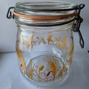 May include: A clear glass storage jar featuring a decorative wrap-around print of golden wheat stalks and small red accents. The jar includes a thick orange rubber gasket seal and a dark metal wire bail closure mechanism secured at the top rim.