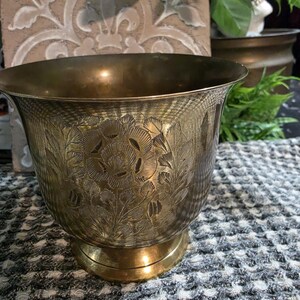 May include: A brass planter with a wide, flared rim and a pedestal base. The exterior surface features an intricate, etched floral design with detailed petals and leaves covering the body of the vessel, showcasing a metallic gold finish.