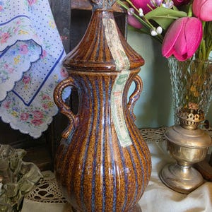 May include: A brown ceramic decorative vase with a lid and two handles. The vase has vertical ribbed detailing with blue accents. It has a label with text on it. The vase is on a table with other decorative items.