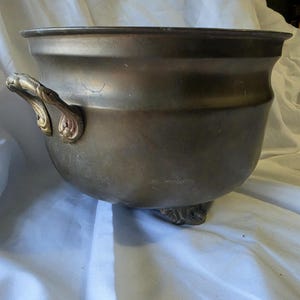 May include: A round, dark metallic bowl with a wide rim and a smooth, weathered surface. It features two ornate, curved handles on the sides and rests on small, decorative feet. The metal has a muted, aged patina with subtle color variations.