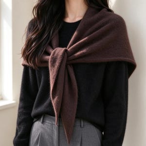 May include: A dark brown, soft-textured triangular scarf tied in a knot at the front, worn over a black long-sleeved sweater and grey trousers. The scarf features a fuzzy, knit appearance and drapes over the shoulders and chest area.