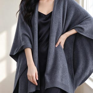 May include: A dark gray open-front poncho with a textured knit finish and ribbed trim along the edges. The garment features wide, loose sleeves and a draped silhouette, worn over a simple black sleeveless slip dress with a V-neckline.