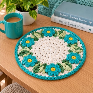 May include: A round, crocheted coaster featuring a cream-colored center surrounded by a border of teal flowers with yellow centers and light green leaves. The coaster has a thick teal braided edge and sits on a wooden surface.