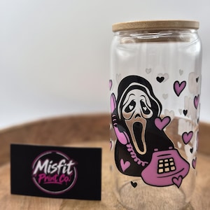 May include: A clear glass tumbler with a bamboo lid featuring a graphic of a masked figure holding a pink rotary telephone, surrounded by small pink and black hearts. A black rectangular card with the text Misfit Print Co. is placed nearby.