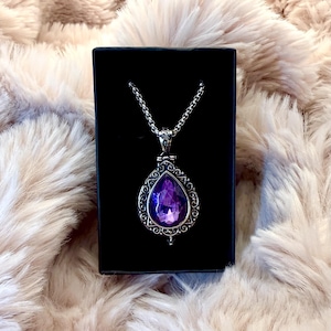 Può includere: Una collana con pendente color argento caratterizzata da una gemma viola a forma di goccia, incastonata in una cornice di metallo decorata con motivi in filigrana. Il pendente pende da una catenina argentata ed è riposto in una scatola nera.