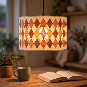 Op de afbeelding: Een cilindervormige hanglampenkap met een herhalend ruitpatroon in tinten oranje, geel en bruin. De kap is gemaakt van een getextureerd, gebroken wit materiaal met doorschijnende, ruitvormige uitsparingen die licht doorlaten.