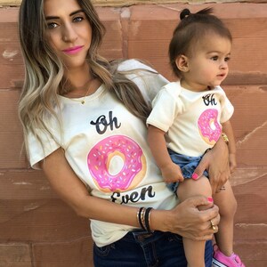 May include: A mother and daughter wearing matching cream colored t-shirts with a pink donut graphic and the text "Oh Even". The daughter is wearing pink sneakers and denim shorts. The mother is wearing blue jeans.