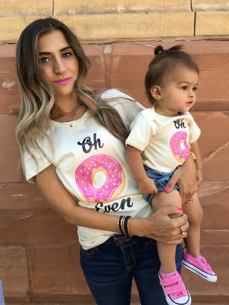 May include: A mother and daughter wearing matching cream colored t-shirts with a pink donut graphic and the text "Oh Even". The daughter is wearing pink sneakers and denim shorts. The mother is wearing blue jeans.
