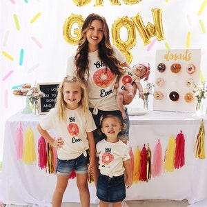 May include: A family of four wearing matching shirts with a donut graphic and the text "Oh Even" on them. The shirts are light beige and the donuts are pink with white frosting. The family is standing in front of a white backdrop with gold foil balloons that spell out "Donut Grow". There is a white sign with the word "donuts" on it and a donut wall behind the family.