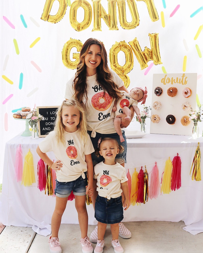 May include: A family of four wearing matching shirts with a donut graphic and the text "Oh Even" on them. The shirts are light beige and the donuts are pink with white frosting. The family is standing in front of a white backdrop with gold foil balloons that spell out "Donut Grow". There is a white sign with the word "donuts" on it and a donut wall behind the family.