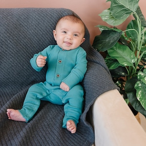 May include: A baby wearing a teal blue long-sleeved onesie with button closures sits on a gray knitted blanket on a white couch. The baby is smiling and looking at the camera.