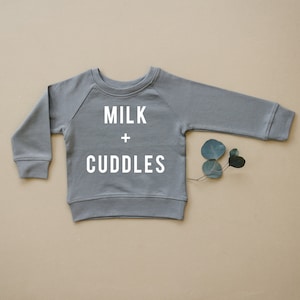Pode incluir: Uma camisa de moletom cinza para bebê com o texto "MILK + CUDDLES" impresso em branco.