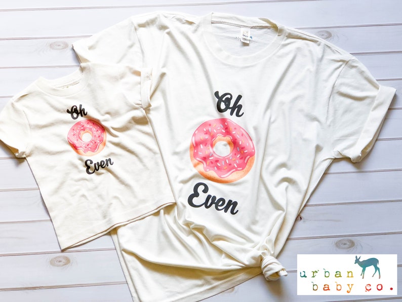 May include: Two cream colored t-shirts with a pink and white donut graphic and the text "Oh Even".