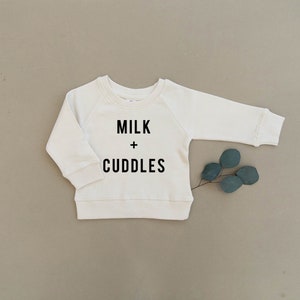 May include: A white long-sleeved sweatshirt with the text "MILK + CUDDLES" printed in black.