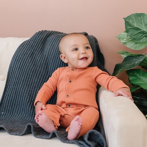 May include: A baby wearing a coral-colored long-sleeved onesie sits on a white couch with a gray knitted blanket. The baby is smiling and looking to the right. A green leafy plant is in the background.
