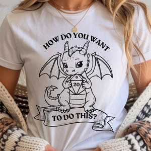 Puede incluir: Una camiseta blanca con un gráfico de líneas negras de un pequeño dragón sosteniendo un dado de veinte caras. Sobre el dragón, el texto dice "HOW DO YOU WANT" y debajo, en una cinta, "TO DO THIS?" en letras mayúsculas negras.