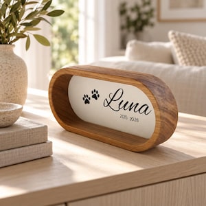 May include: An oval-shaped wooden frame with a white centre panel. The panel features two black paw prints, the name Luna in a cursive script, and the years 2015 and 2026 printed in black text. The frame has a natural wood grain finish.