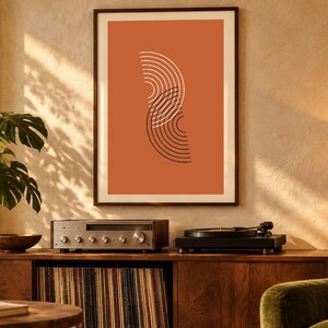 May include: A framed wall art print featuring a burnt orange background with two overlapping vertical semi-circles composed of thin white and black concentric lines, displayed in a dark wood frame on a textured wall.