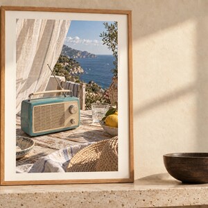 May include: A framed wall art print featuring a teal vintage-style radio with a cream-coloured speaker grille and handle, sitting on a weathered wooden table. The scene includes a straw hat, a bowl of lemons, and a glass, with a coastal view in the background.