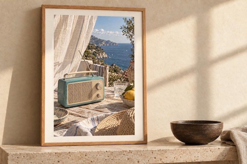 May include: A framed wall art print featuring a teal vintage-style radio with a cream-coloured speaker grille and handle, sitting on a weathered wooden table. The scene includes a straw hat, a bowl of lemons, and a glass, with a coastal view in the background.