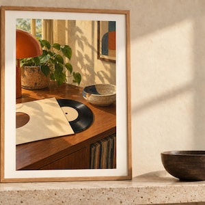 May include: A framed wall art print featuring a warm-toned interior scene with a wooden record player console, a black vinyl record in a beige sleeve, a green potted plant, an orange lamp, and a speckled ceramic bowl on a wooden surface.